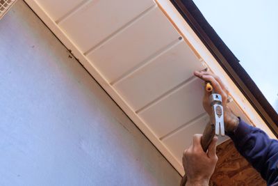 Person hammering siding on a building's eave, beige siding and trim, blue sky.
