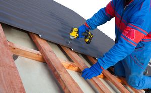 Person in blue overalls installing roofing material with a power drill.