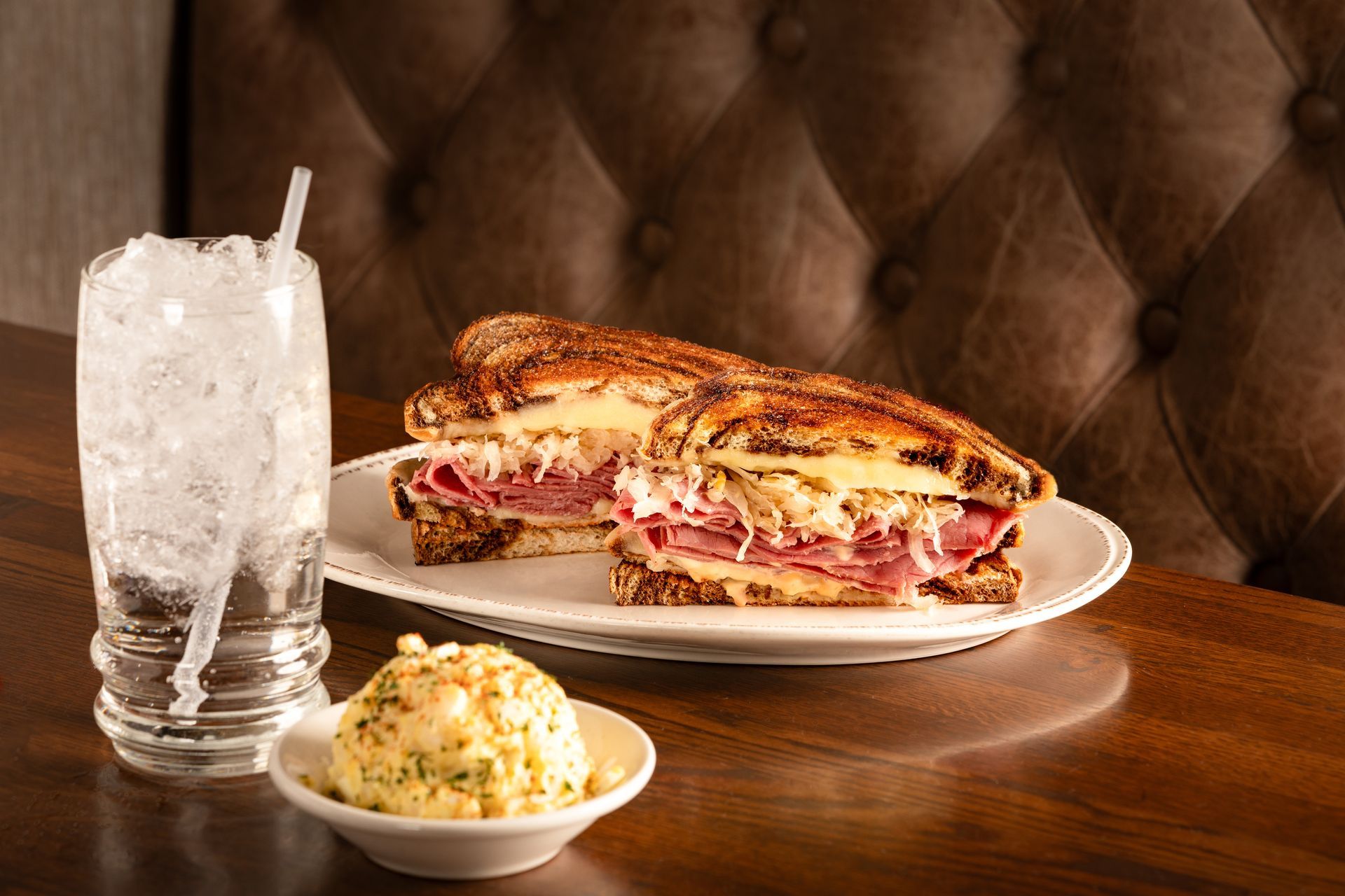 Reuben sandwich and potato salad on a plate, next to a glass of ice water on a dark wood table.