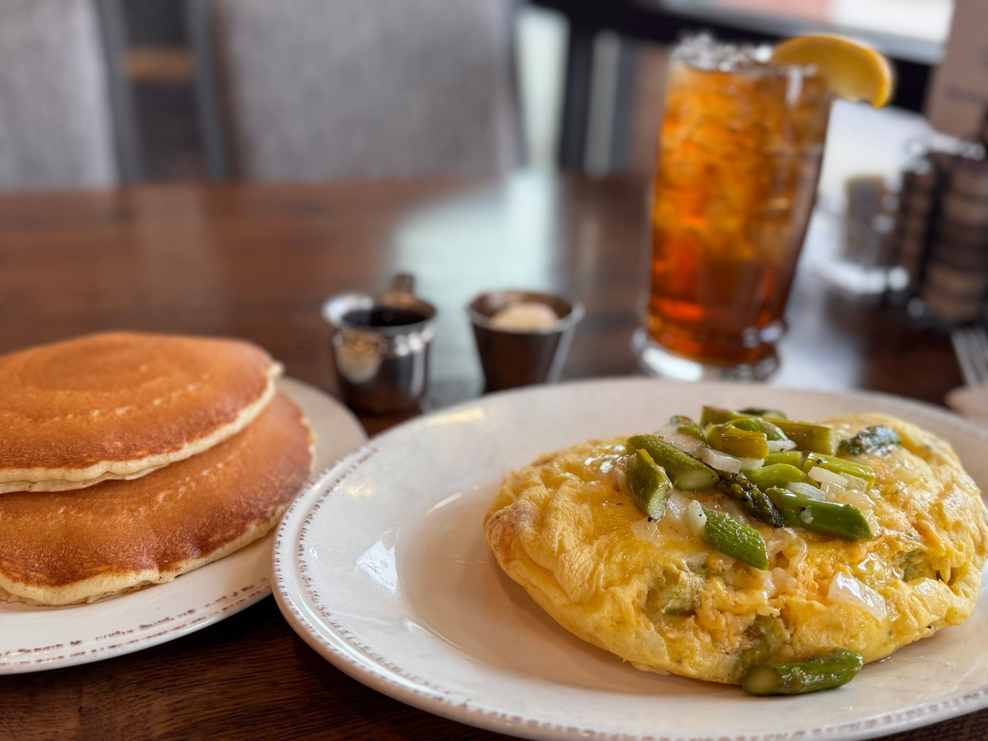 Pancakes and omelet topped with asparagus on a table with iced tea, syrup, and butter.