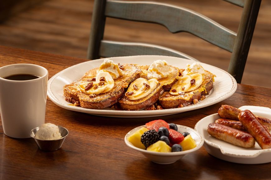 Breakfast: French toast with bananas, fruit, sausage, coffee, and butter on a wooden table.