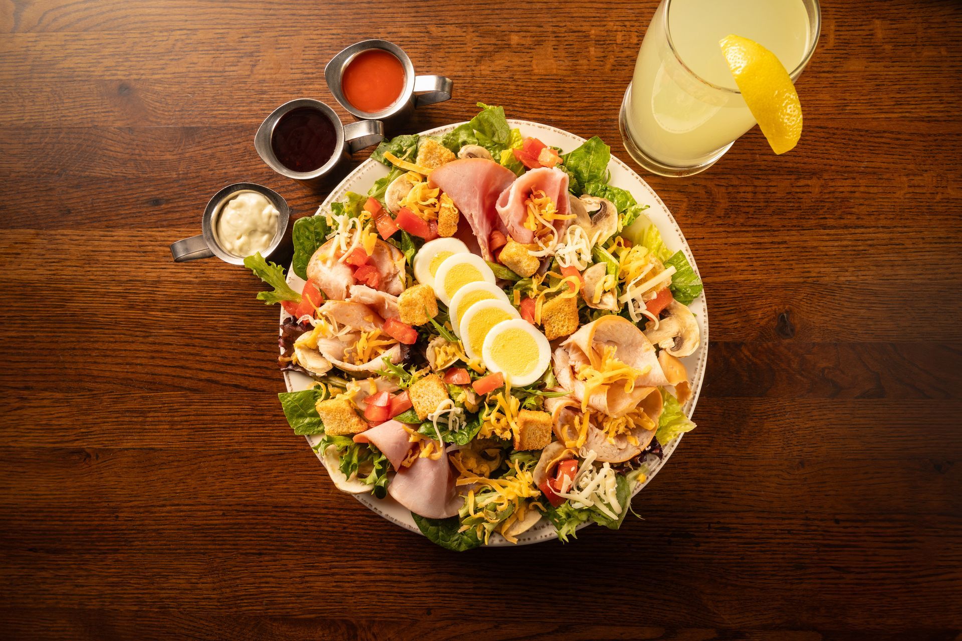 Salad with eggs, ham, cheese, and dressing, alongside lemonade on a wooden table.
