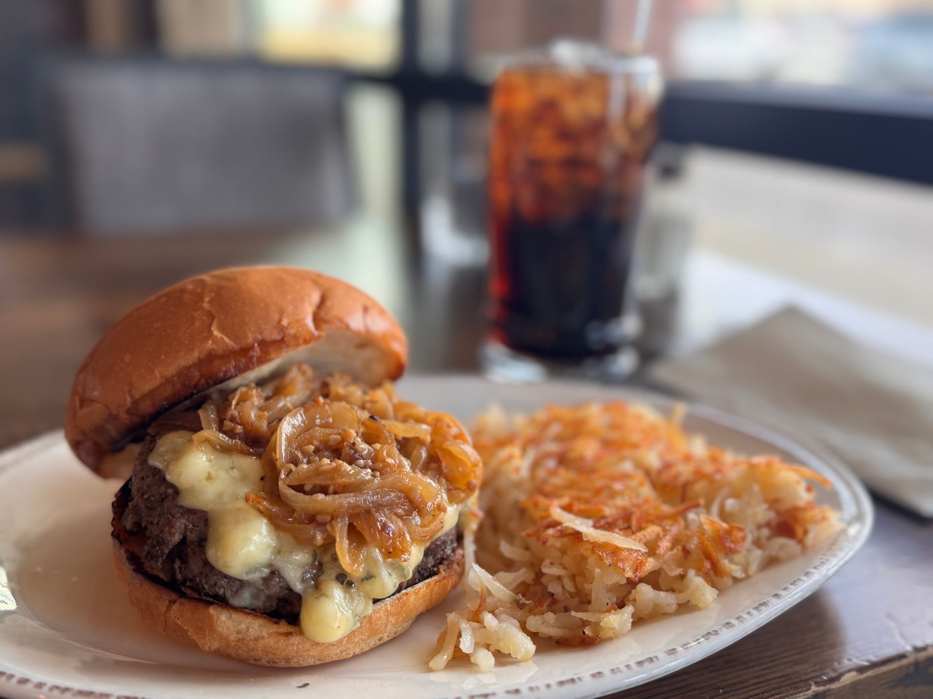 Burger with caramelized onions and hash browns, alongside a glass of soda.