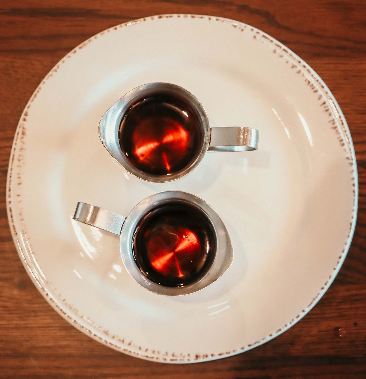 Two small metal pitchers of dark liquid on a white plate with a rustic edge, set on a wooden surface.