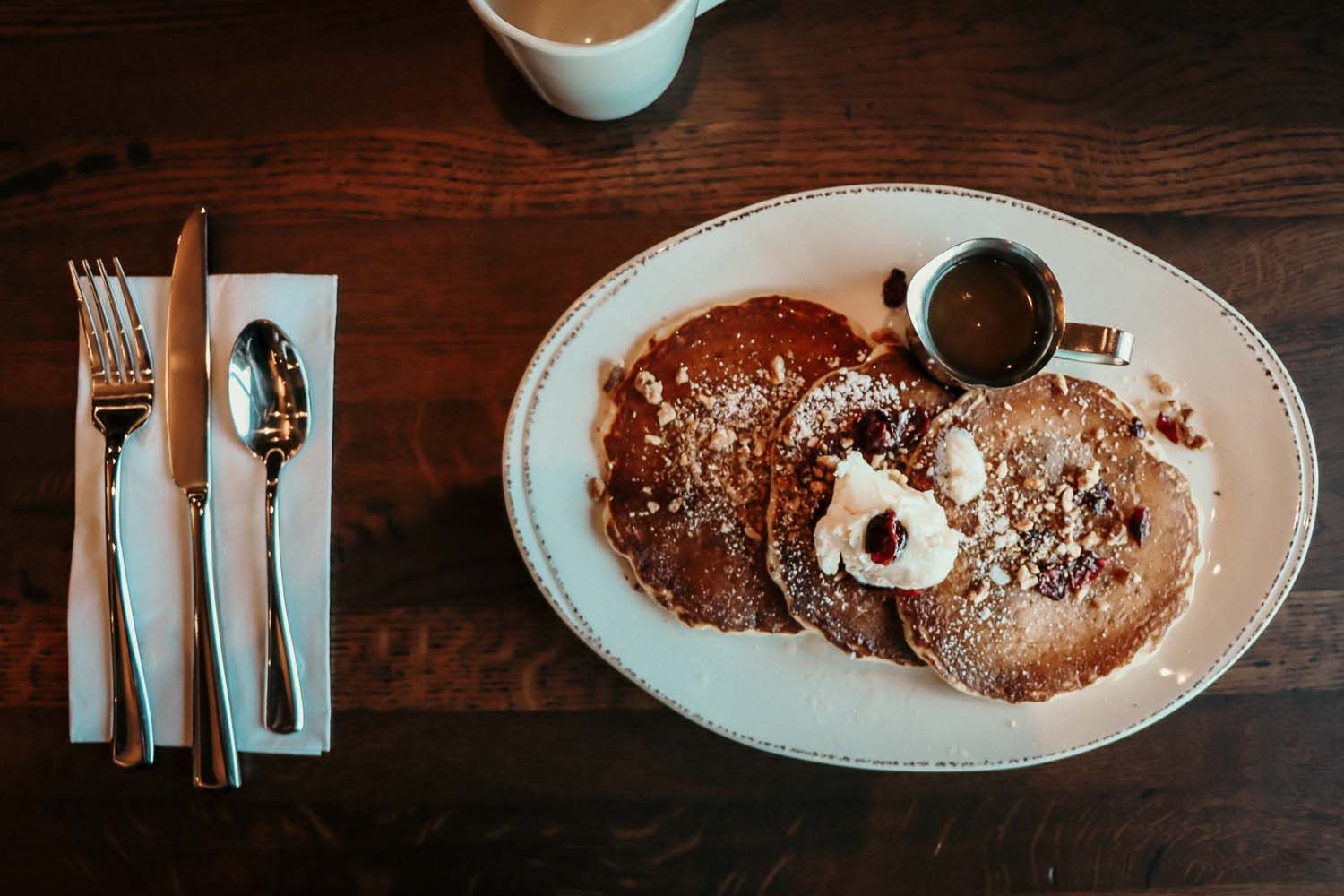 Pancakes on a plate with syrup and butter, silverware and a coffee cup on a wooden table.
