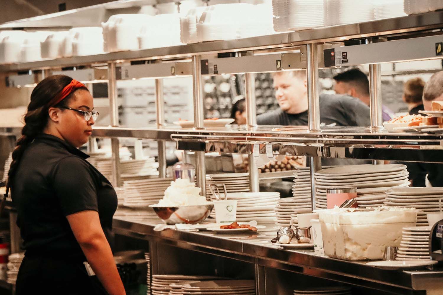 Woman with glasses in a busy restaurant kitchen, holding a plate of food, looking towards the food.