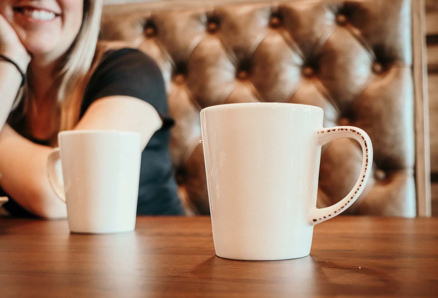 Two white coffee mugs on a brown table with a smiling person in the background, near a tufted leather booth.