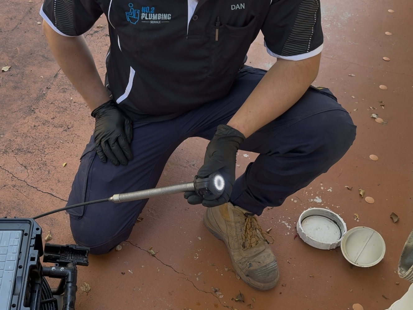 Person in work attire inspecting a surface with a tool, outdoors, with containers of substance visible. — No.2 Plumbing In Kenmore, QLD