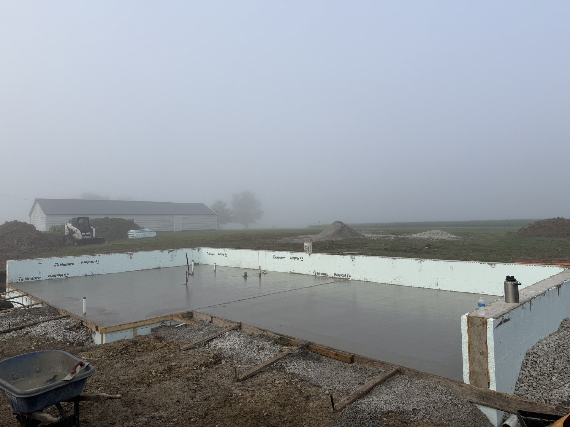 Foggy outdoor construction site with concrete foundation walls and a wheelbarrow in the foreground