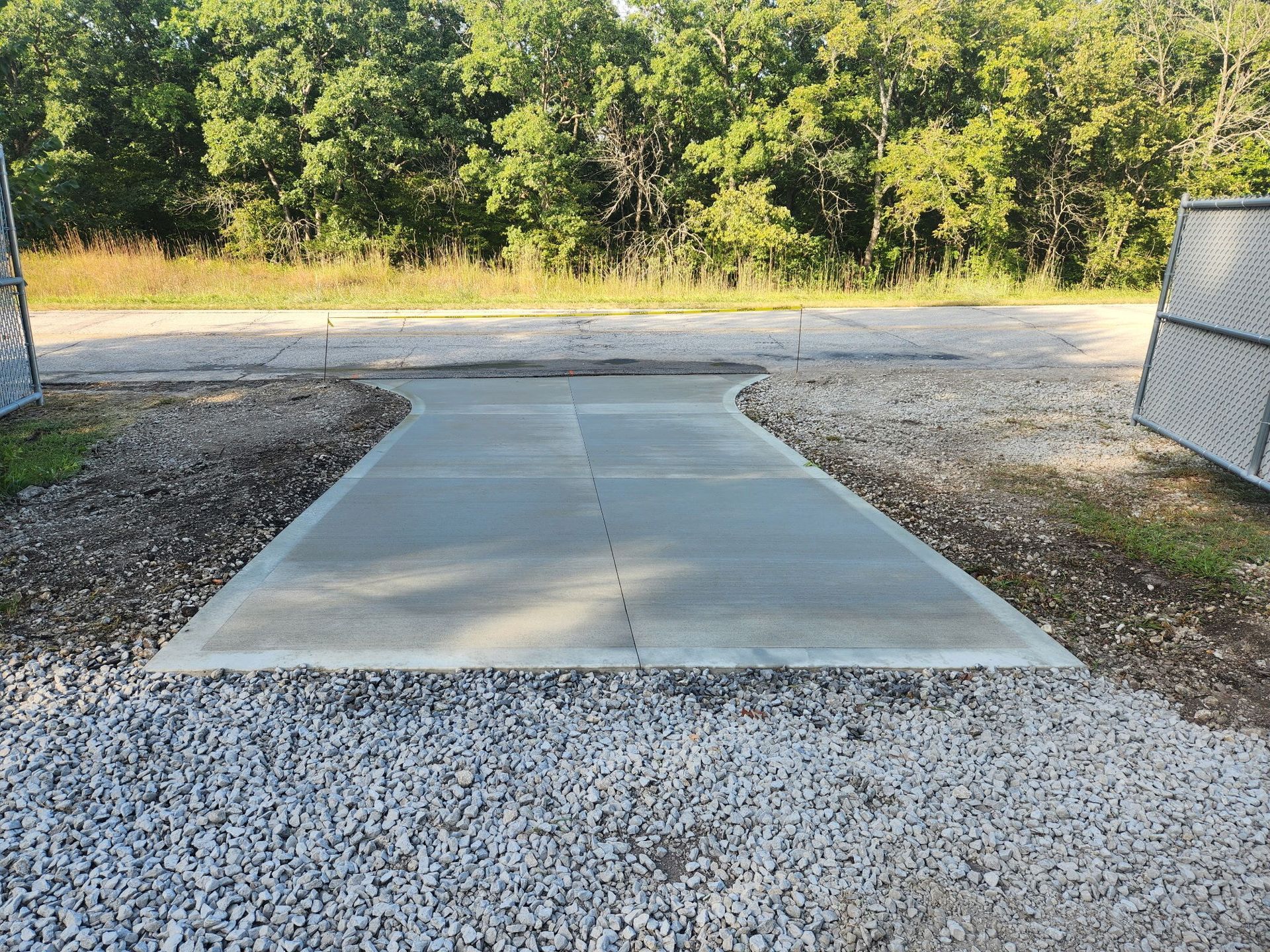 Concrete walkway leading to a gravel path, bordered by grass and trees in a wooded area.