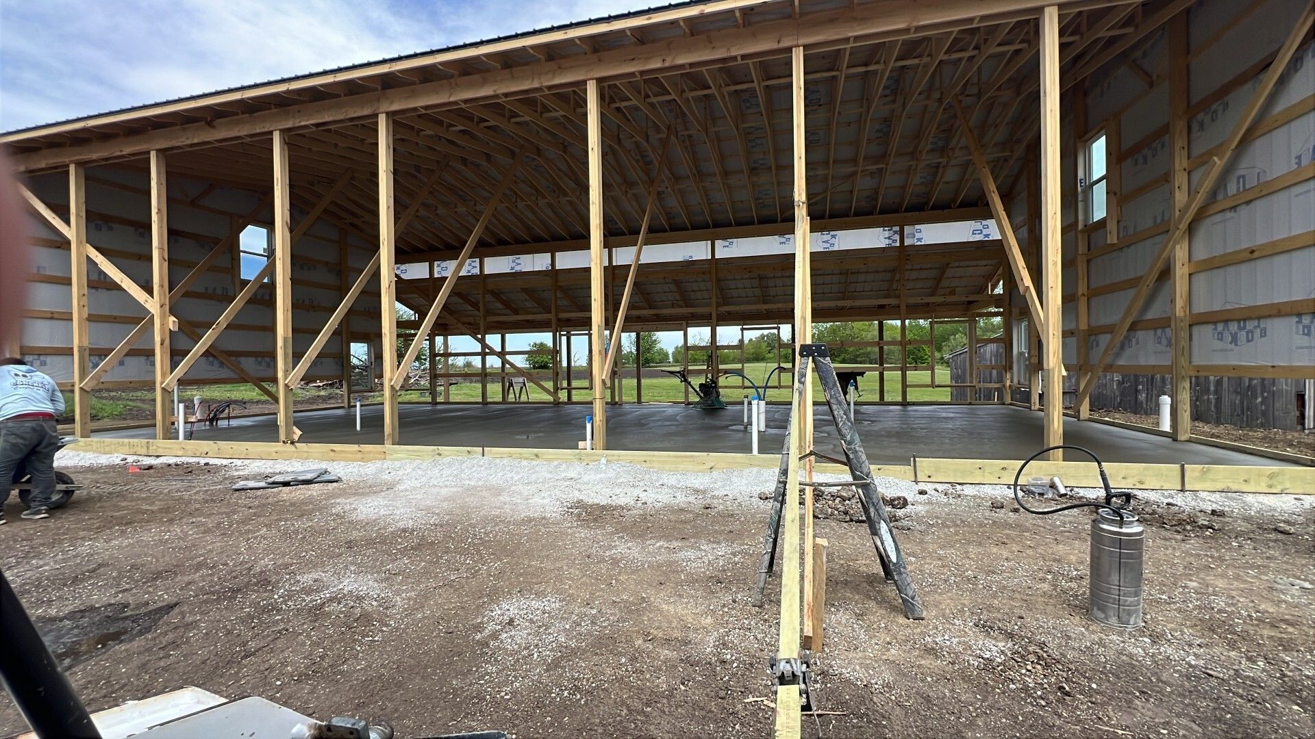 Partially built wooden barn with open framing and roof, gravel foreground, and ladder in front
