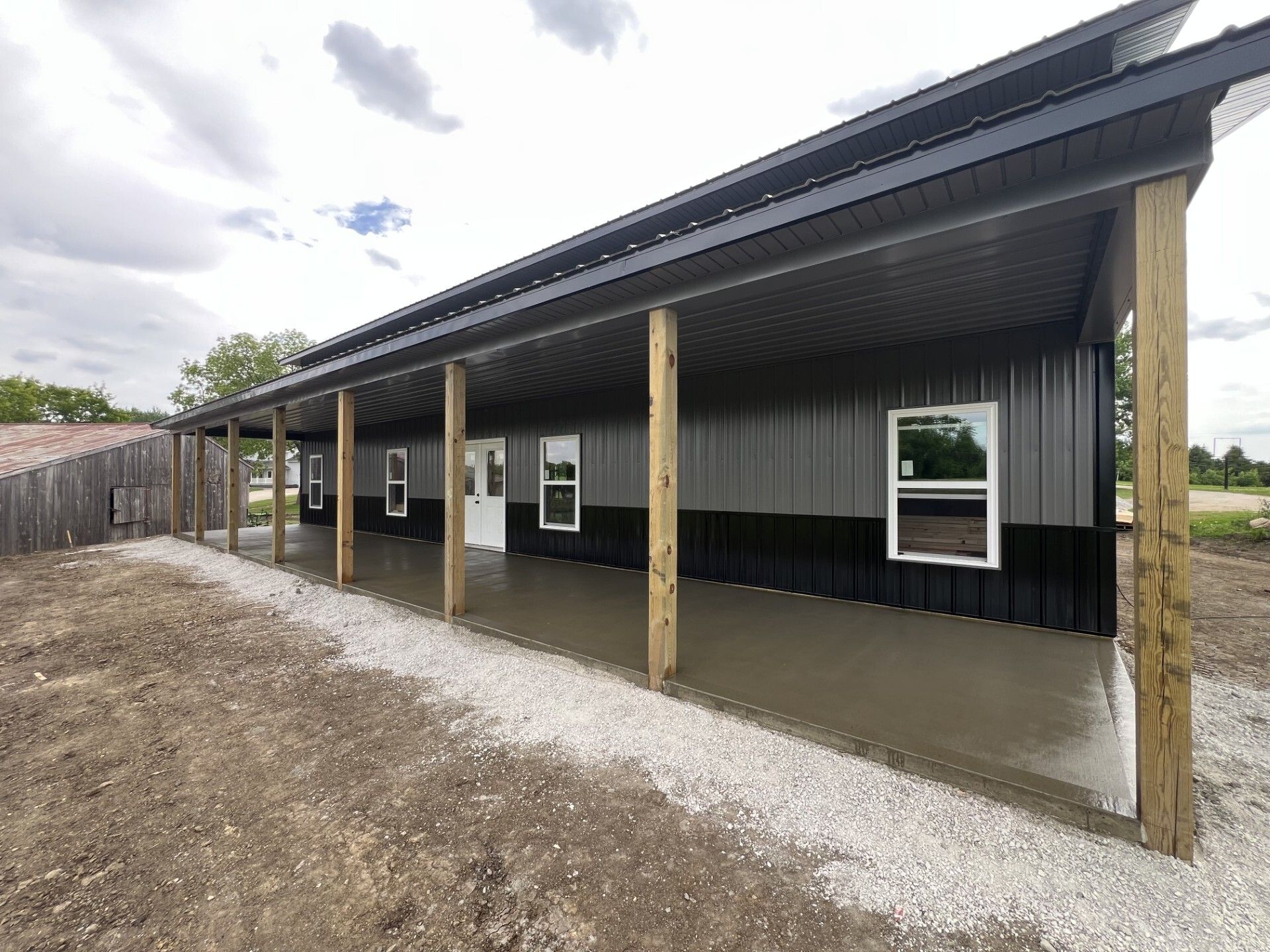 Long, covered porch on a small building with black siding, wood posts, and gravel ground under cloudy sky