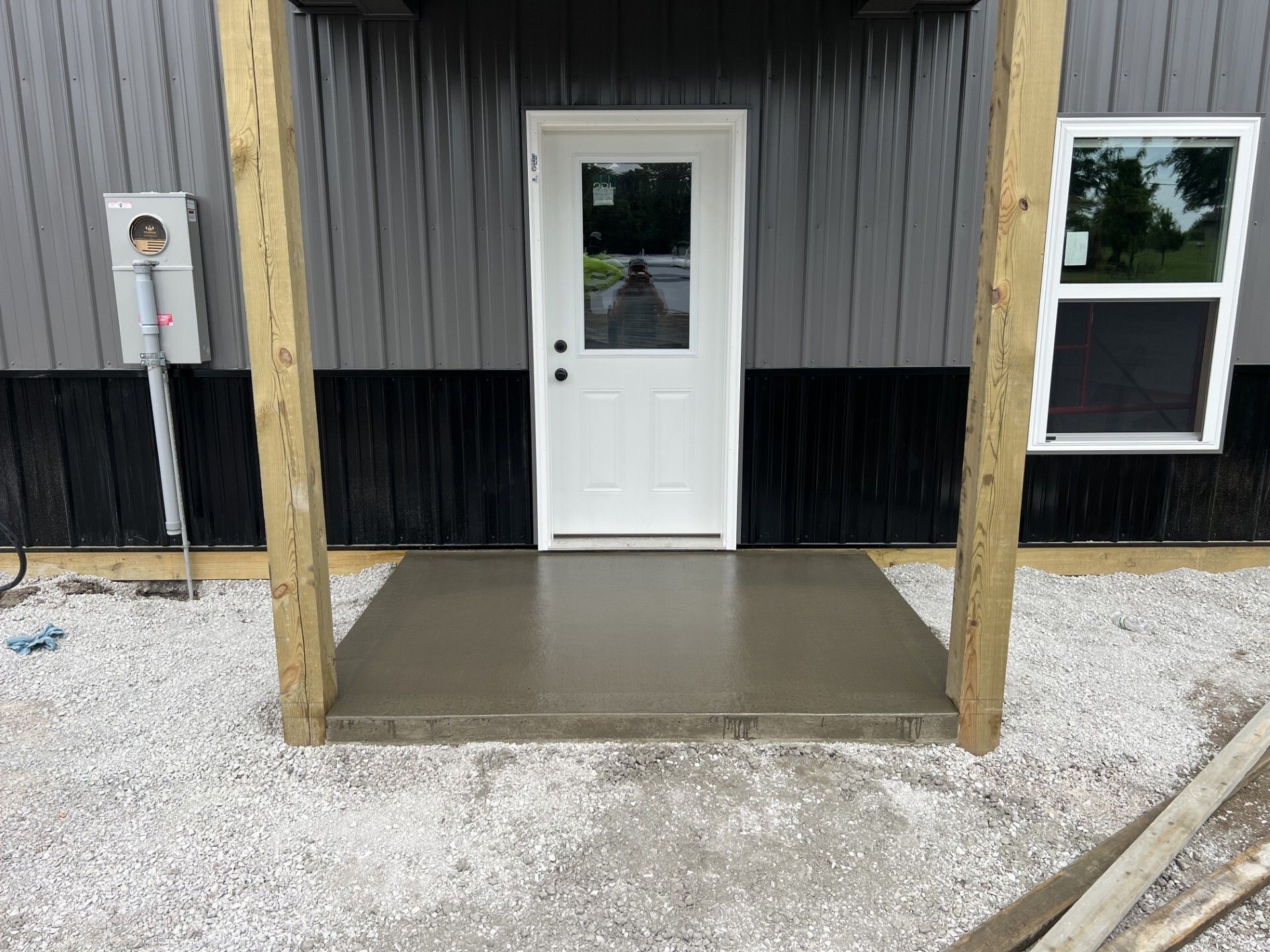 Front entrance with white door, black siding, wooden posts, and a concrete porch over gravel.
