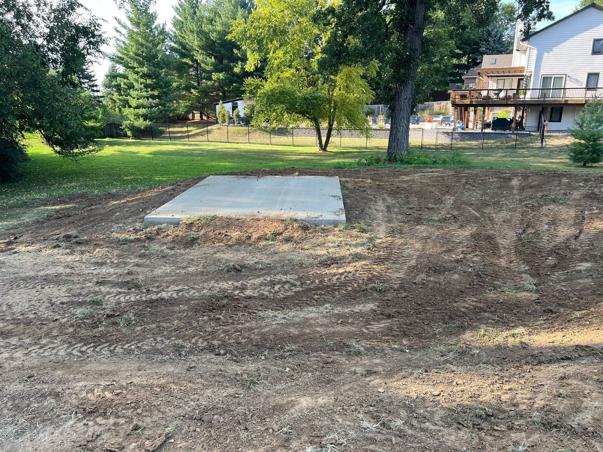 Bare dirt yard with a small concrete slab, trees, and a house in the background