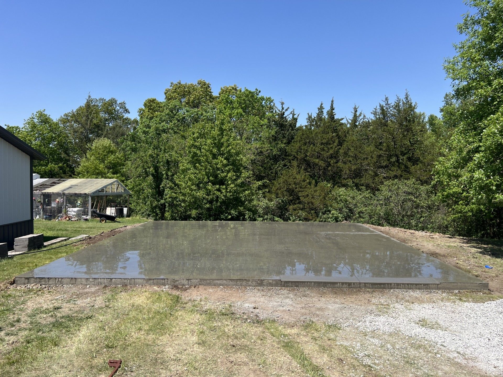 Flooded gravel pad in a sunny backyard, with trees and a small house in the background.