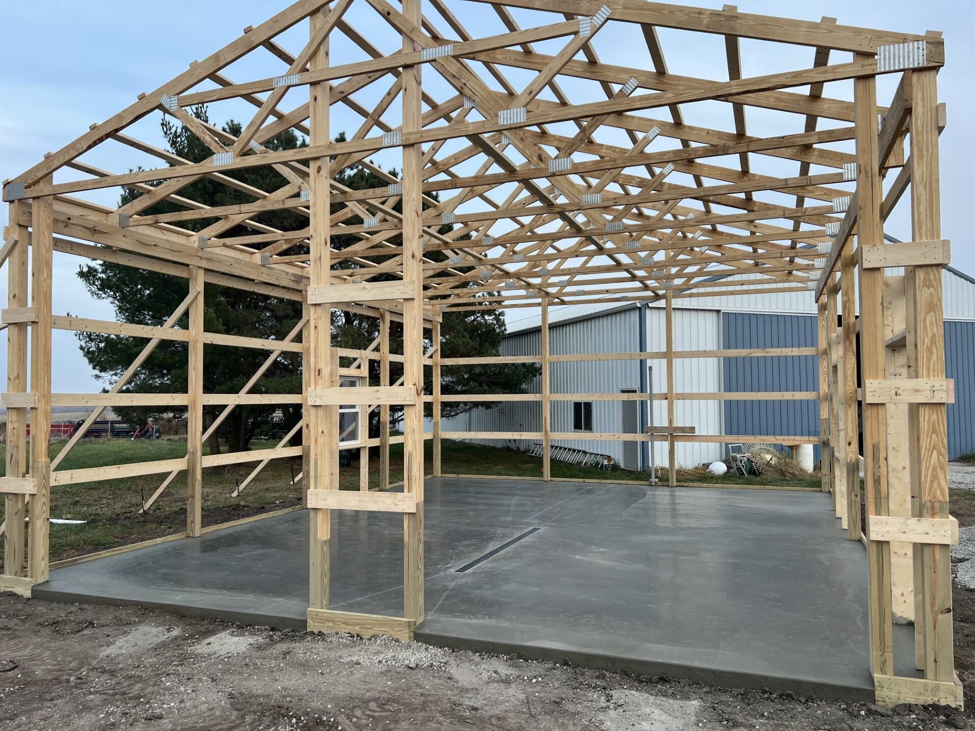 Wooden timber frame structure under construction on a concrete slab beside a metal building