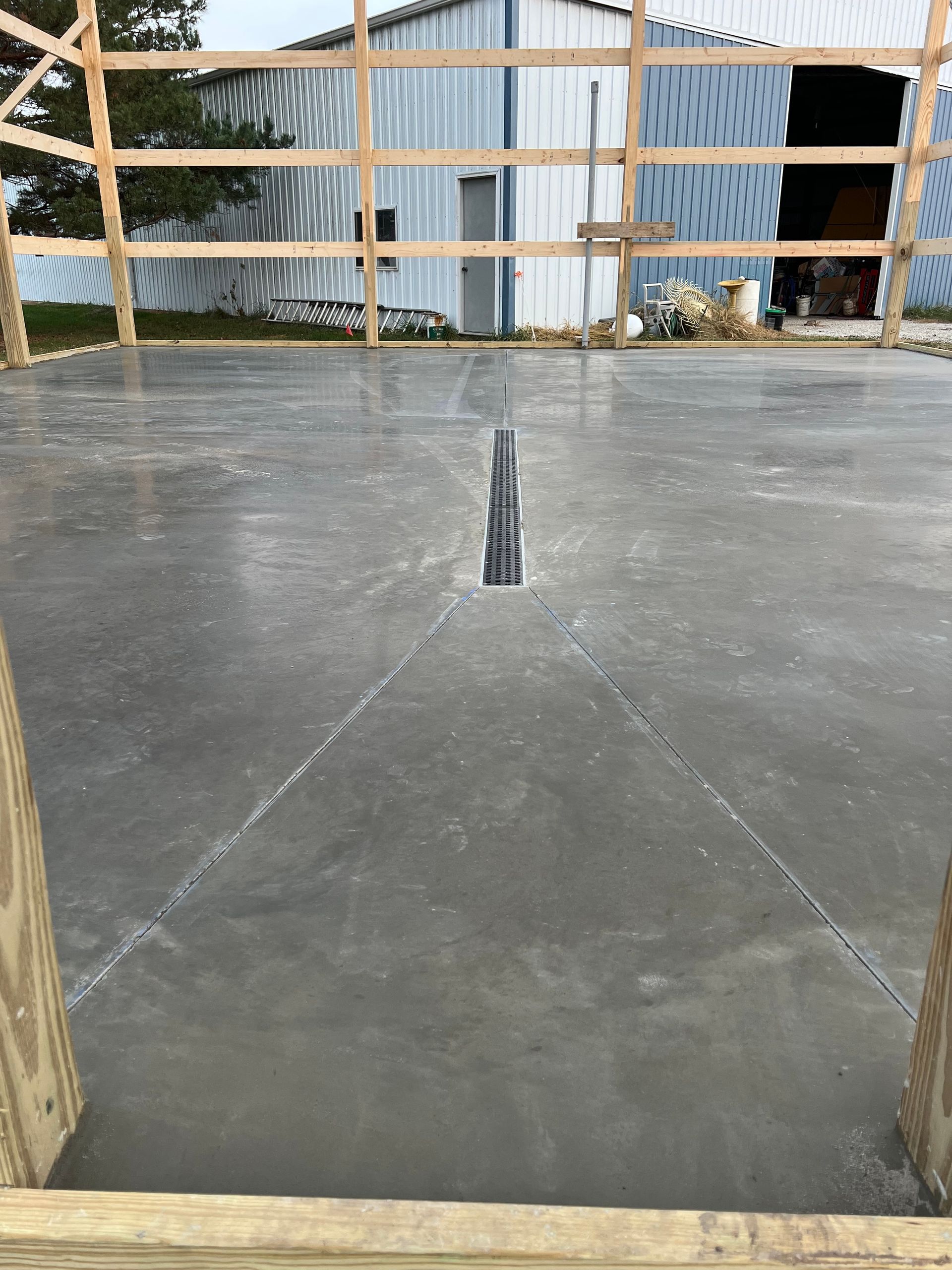 Concrete slab with a narrow center joint inside a partially framed structure, with a barn in the background