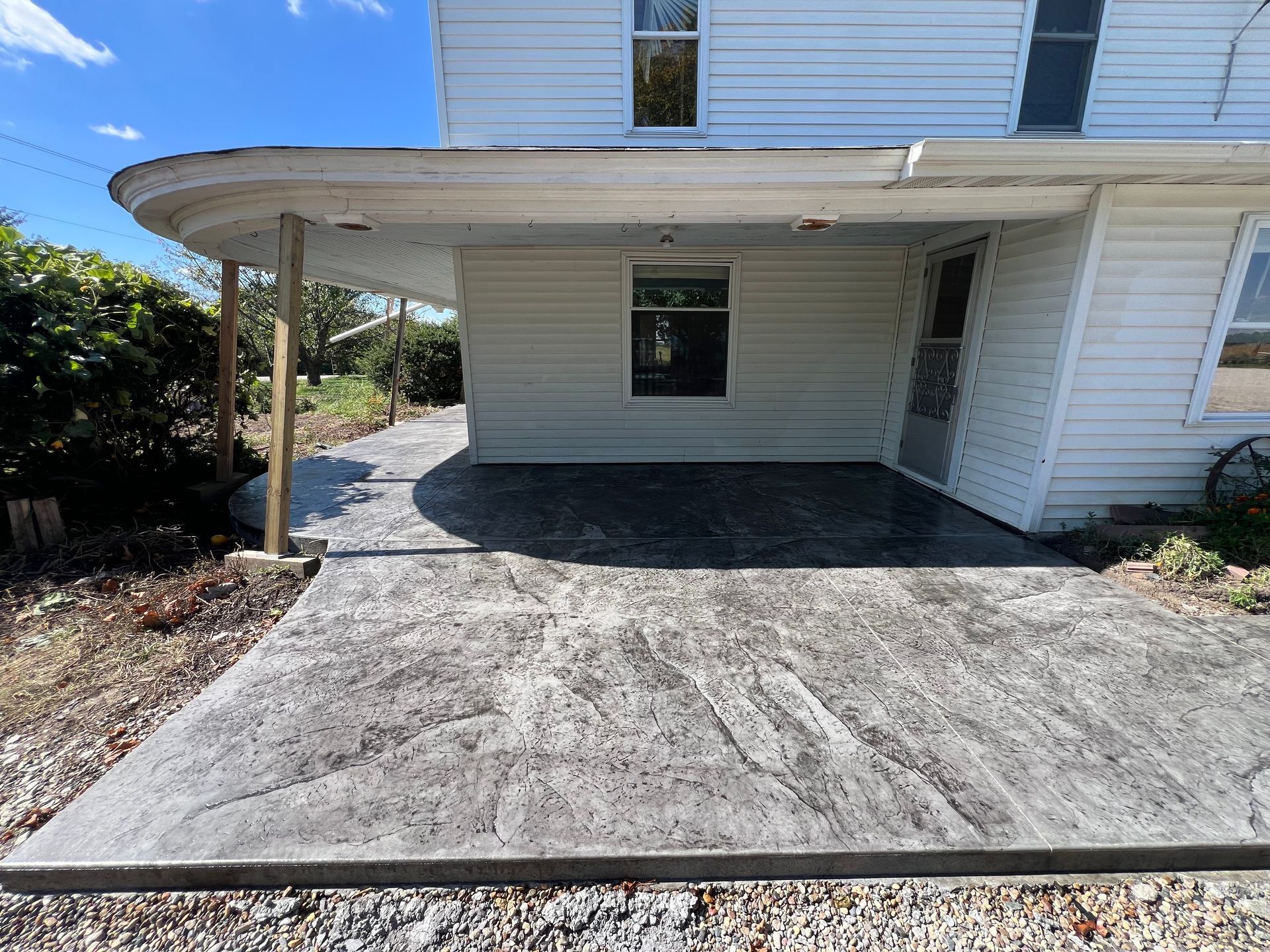 Concrete patio beside a white house with a covered overhang and window under a blue sky