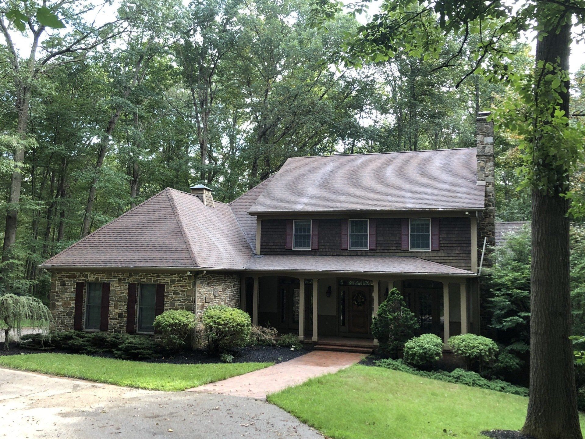 Two-story house with brown roof and porch, surrounded by trees and lawn, connected by a curved walkway.