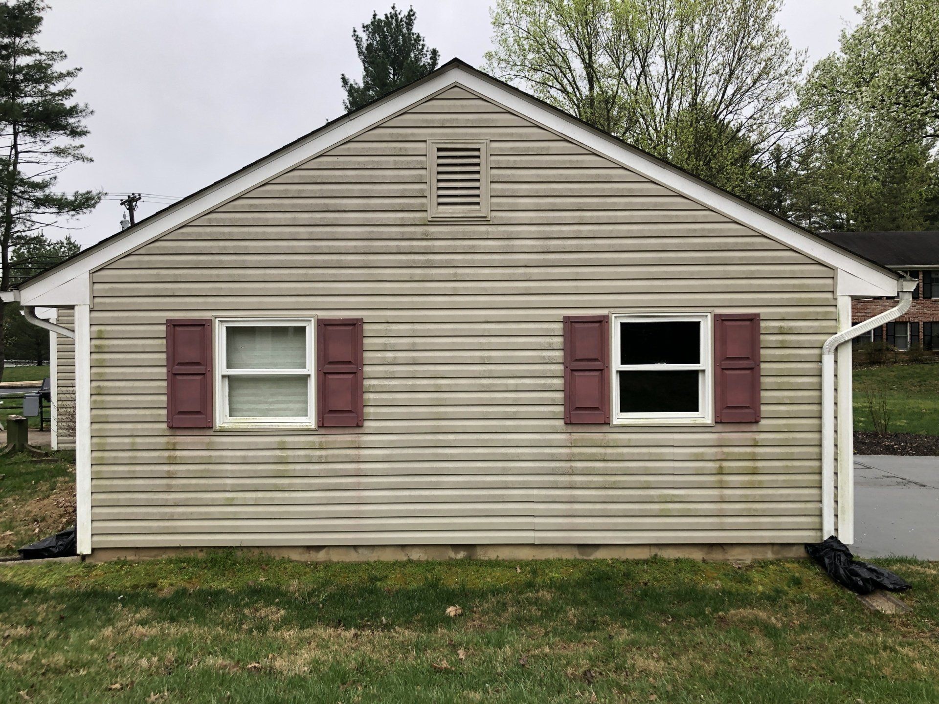 Front of a beige house with maroon shutters and a small attic vent, set against trees and grass.