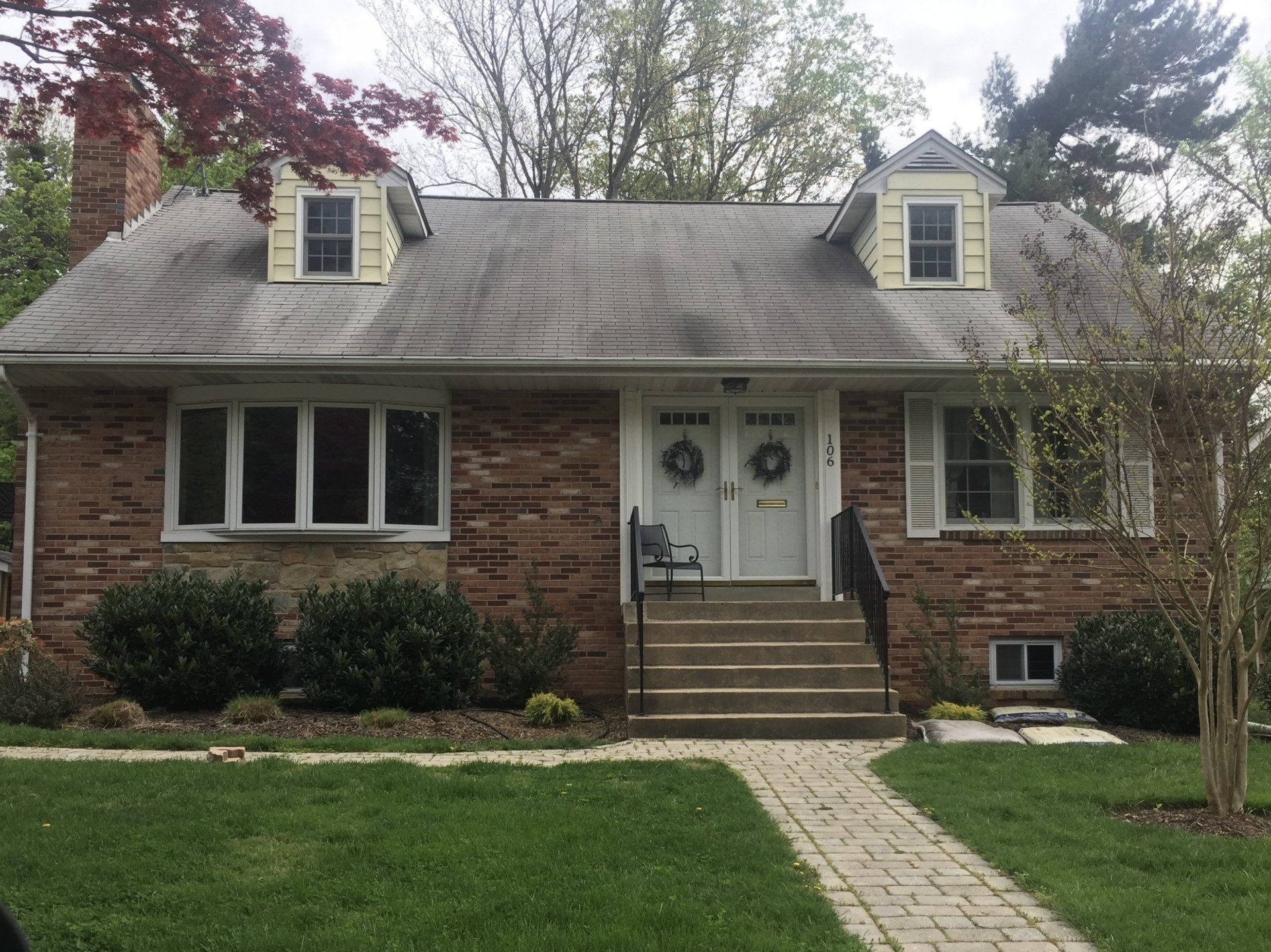 Brick house with dormer windows, front steps, and a central white door framed by shrubs and a lawn.