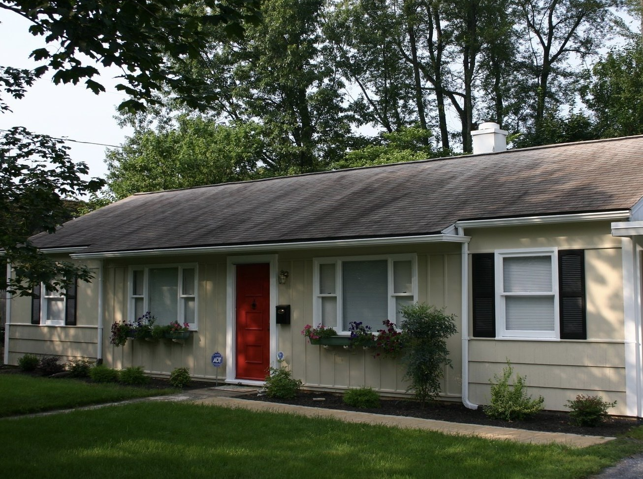 Single-story beige house with a red front door, white shutters, and trees in the yard