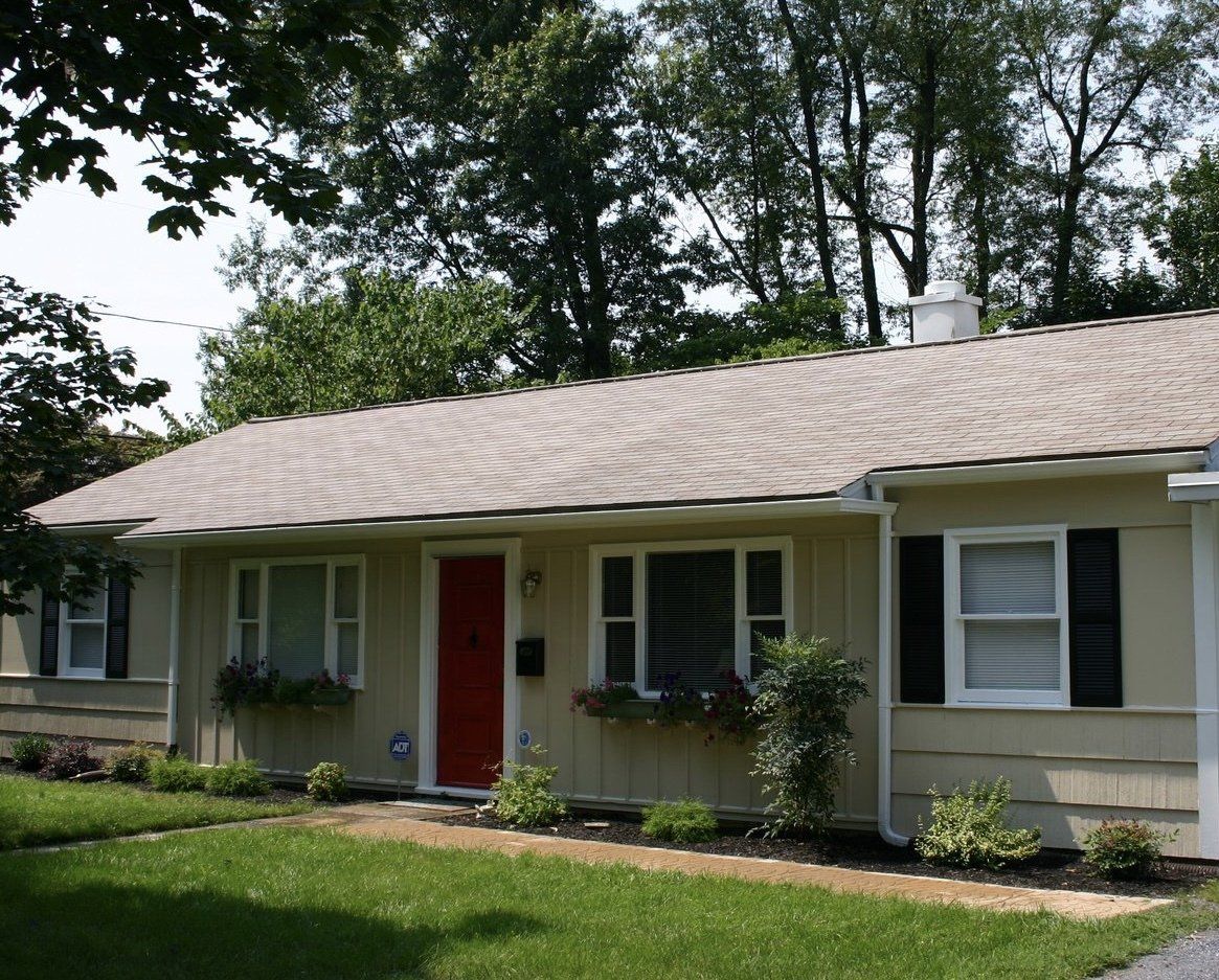 Single-story beige house with a red front door, white shutters, and a green lawn under trees