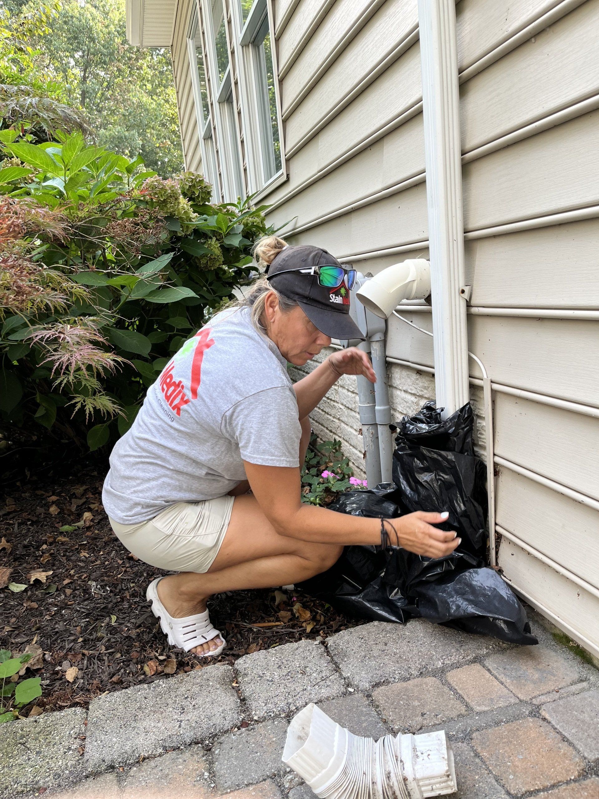 Person crouches by house utility pipes, working beside a brick patio and siding.