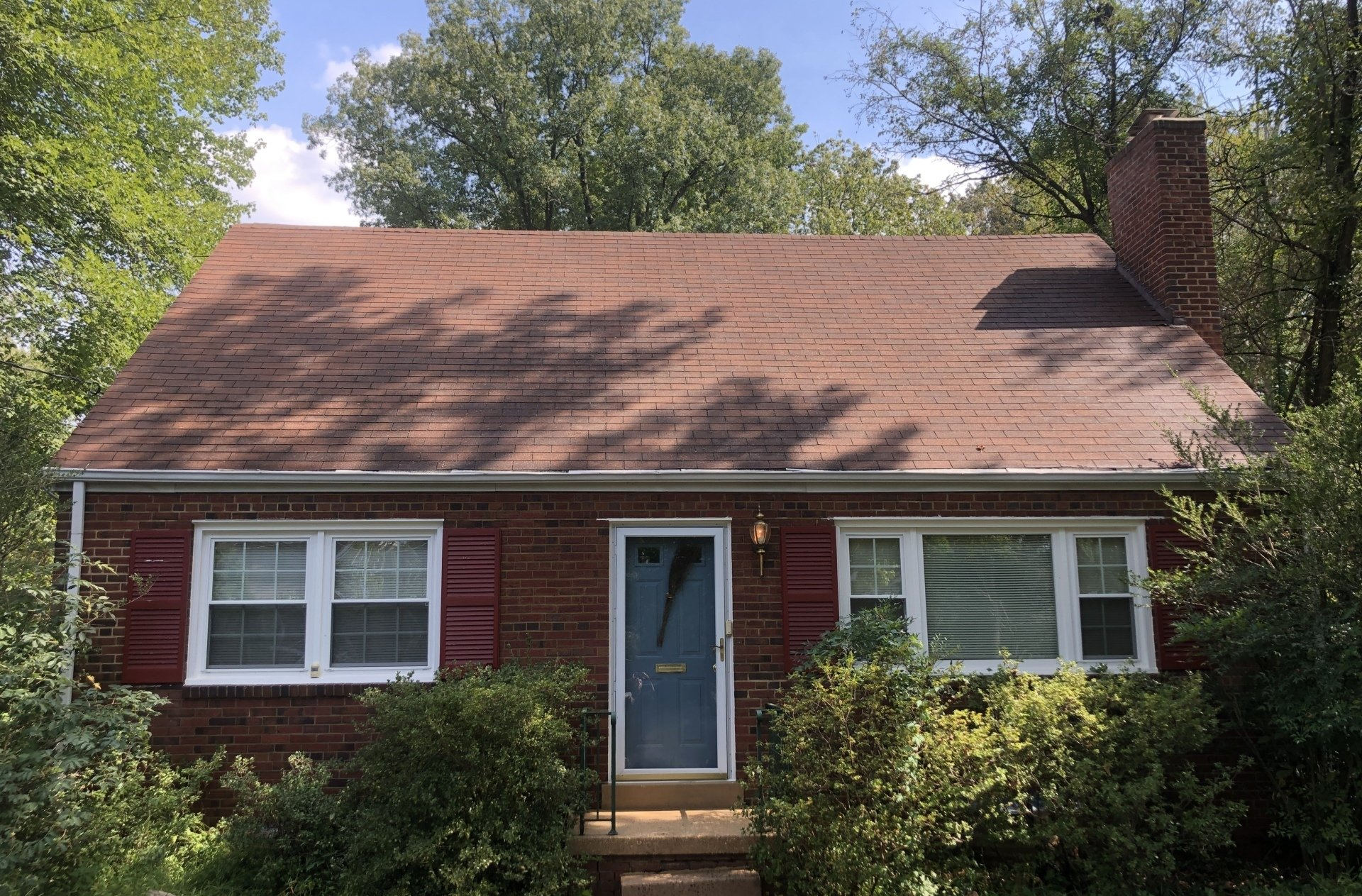 Small red brick house with blue front door, white-trimmed windows, and a shingled roof surrounded by trees