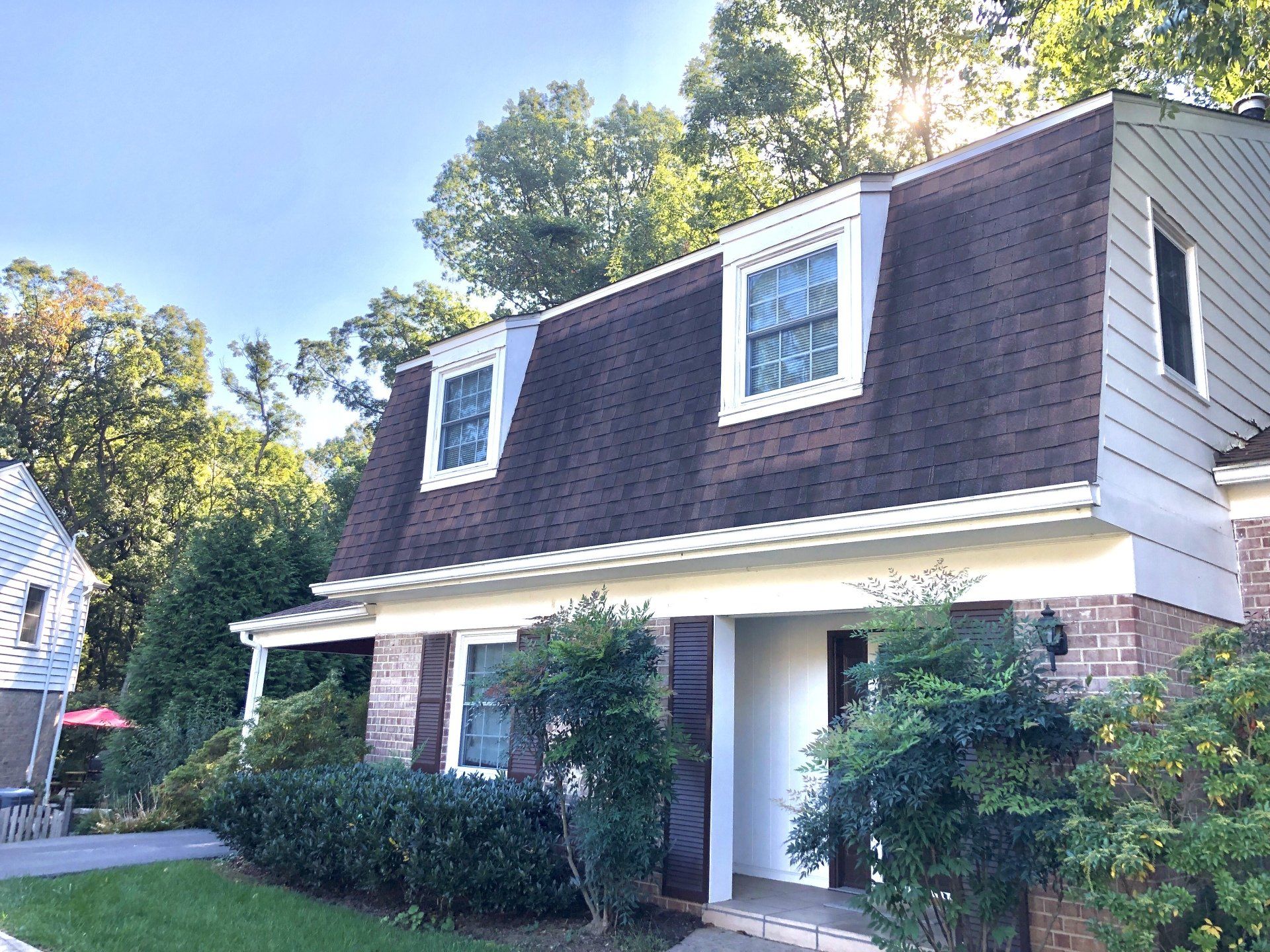 Suburban two-story house with brown shingled roof, brick lower facade, and white trim surrounded by trees.