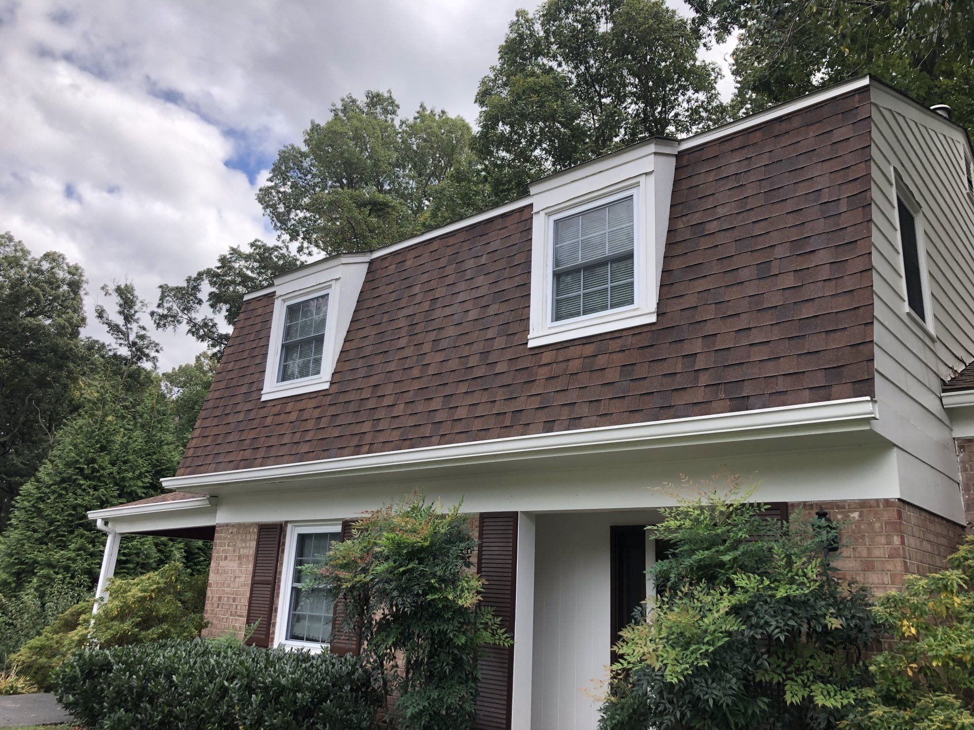 Two-story house with brown shingle roof, white trim, and brick facade surrounded by trees