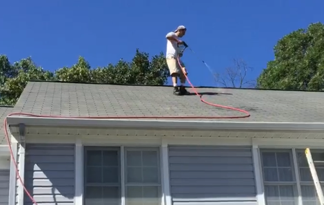 Worker on a roof spraying or cleaning shingles with a hose under a clear blue sky
