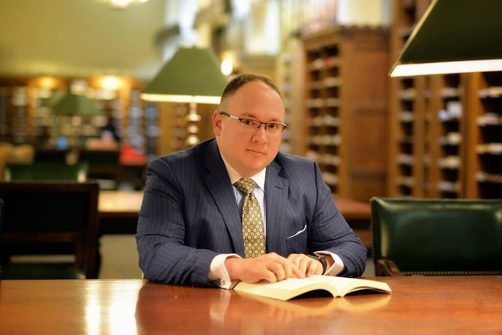 Man in a blue suit reading a book at a wooden table in a softly lit library.
