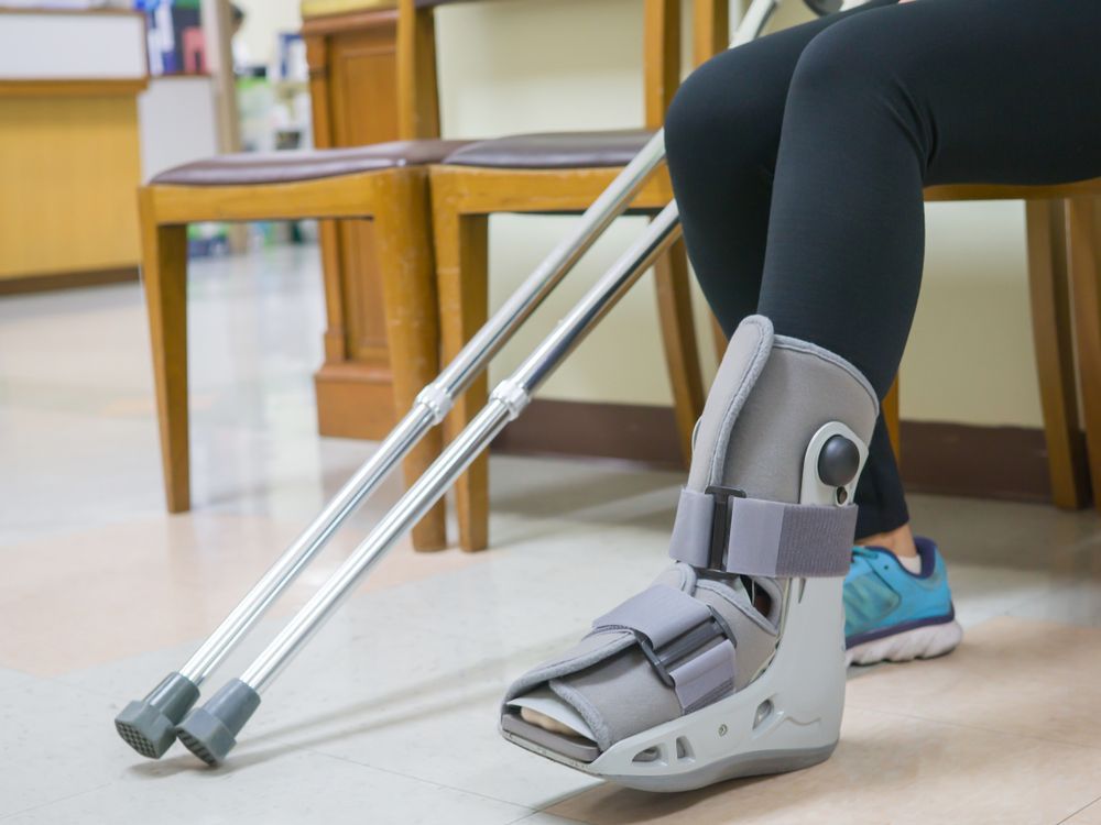 Person wearing a gray ankle brace sits with crutches on a clinic floor.