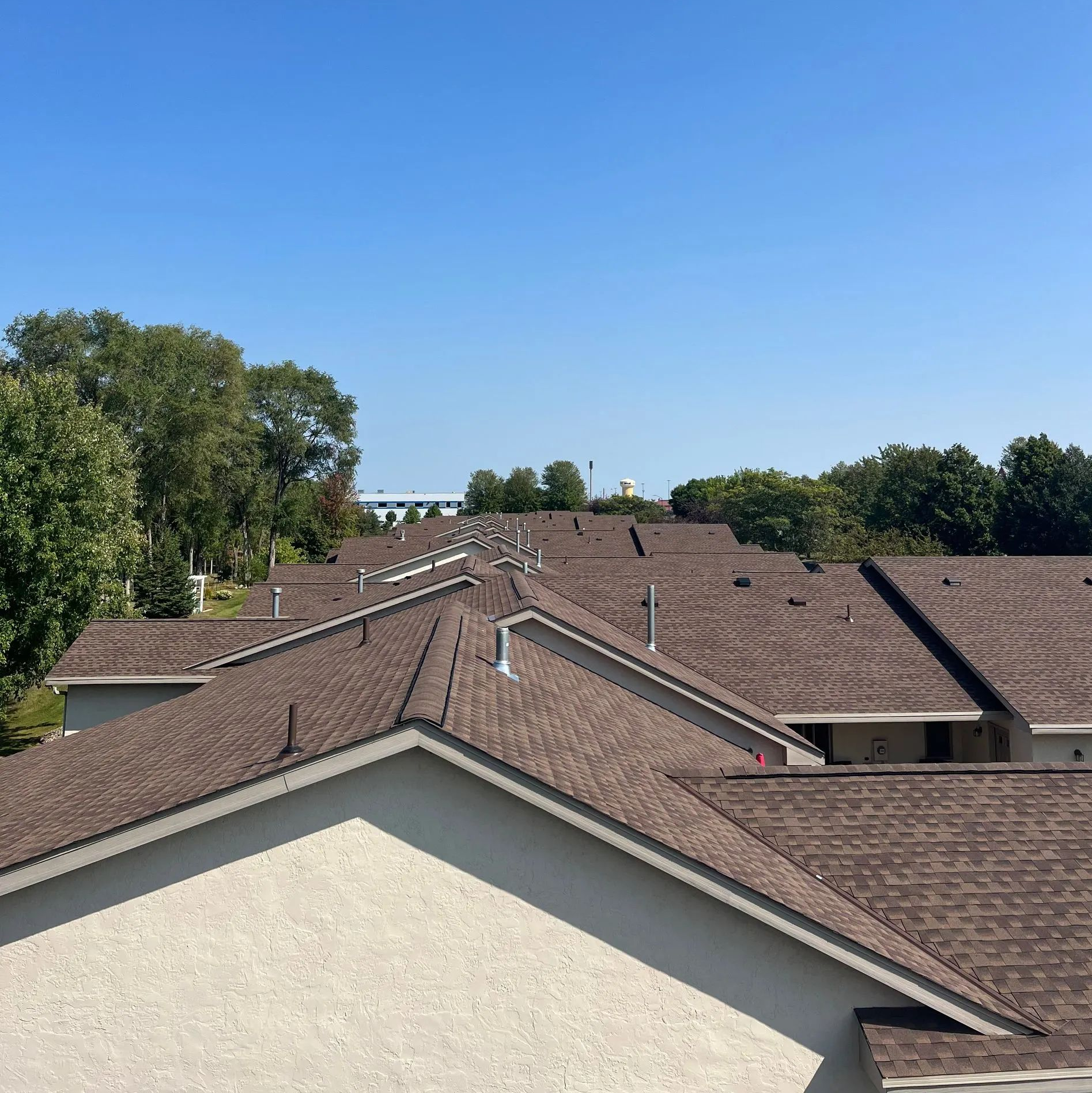 Rows of brown shingled rooftops under a clear blue sky, viewed from above with trees in the distance