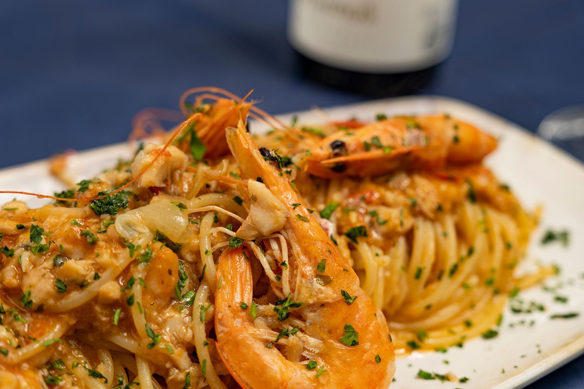 Pasta con gamberi, guarnita con erbe aromatiche, su un piatto bianco; bottiglia di vino sullo sfondo.