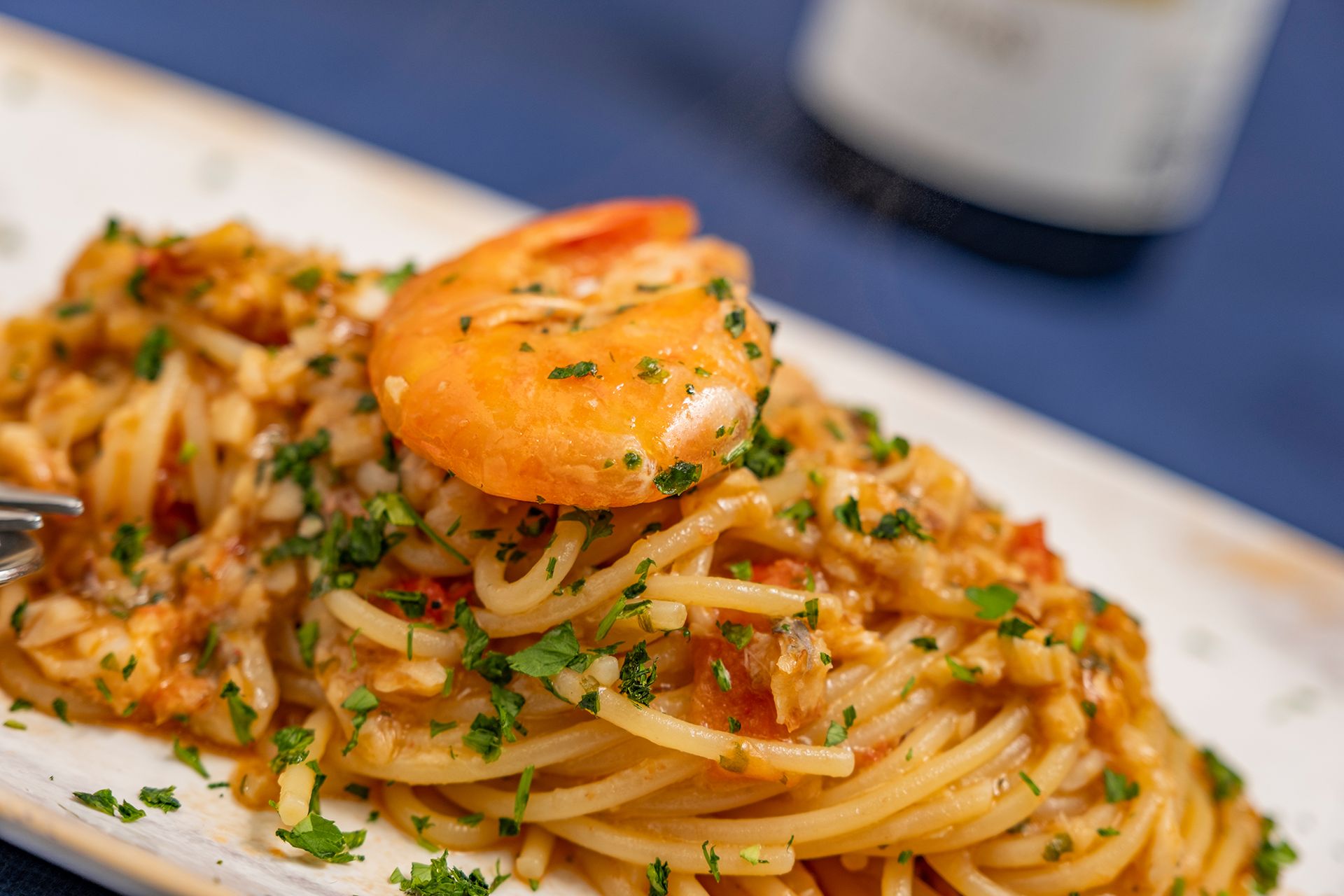 Spaghetti con gamberi e prezzemolo su un piatto bianco, sfondo blu.