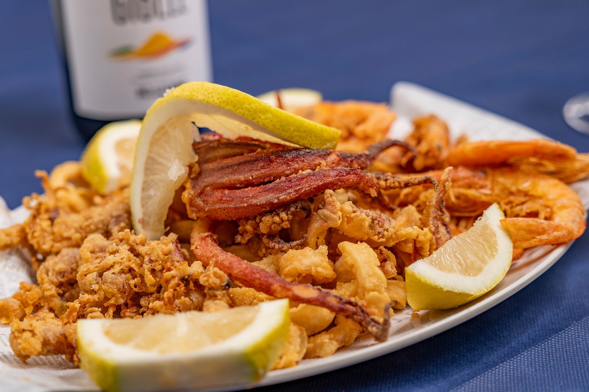 Piatto di pesce fritto con fette di limone su un piatto bianco, sfondo blu. Una bottiglia di vino bianco sullo sfondo.