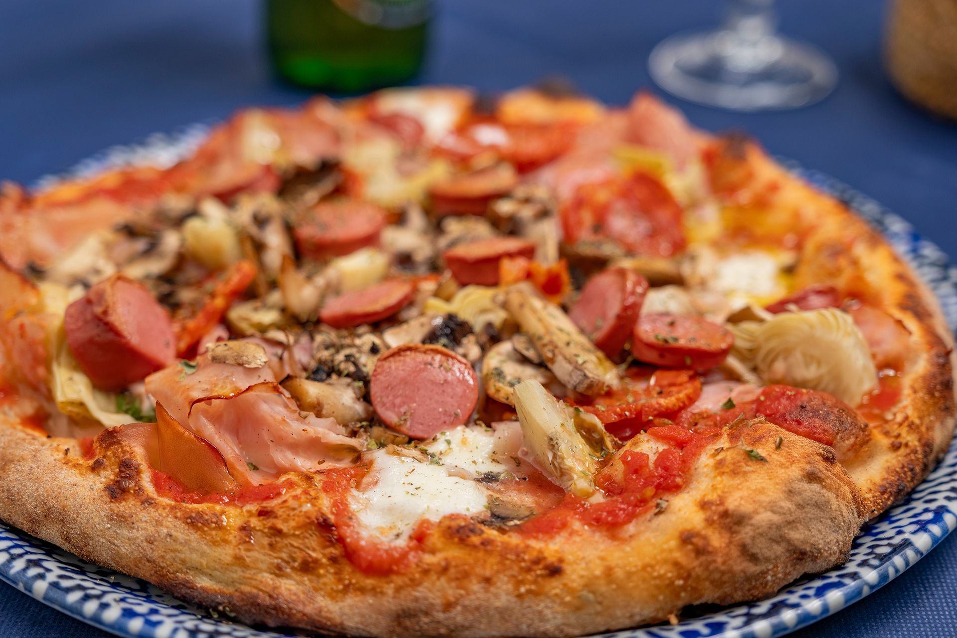 Pizza con salsiccia, prosciutto, funghi e carciofi su un piatto con bordo blu.