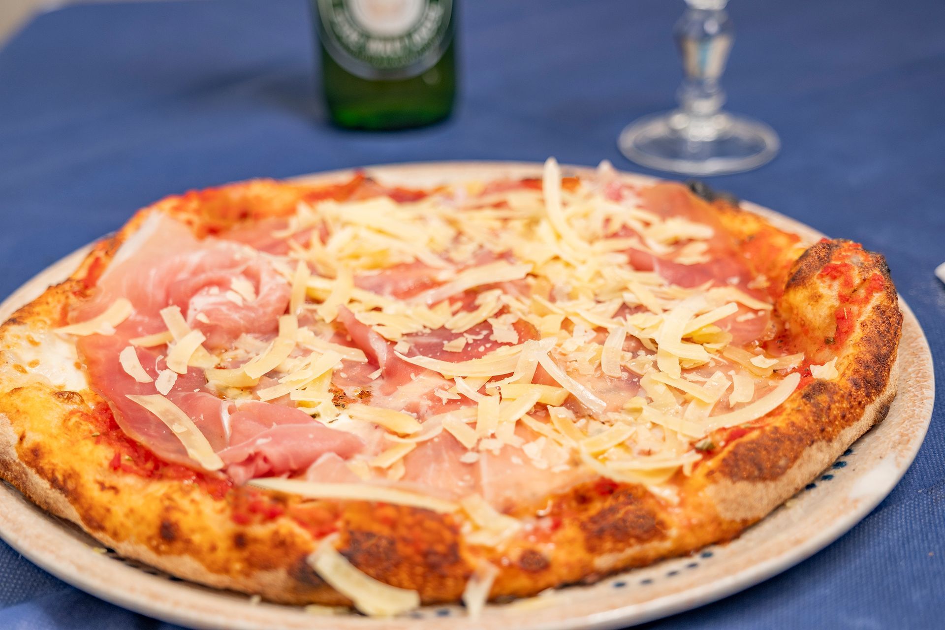 Pizza con prosciutto e formaggio su un piatto, su una tovaglia blu. Bottiglia e bicchiere sfocati sullo sfondo.