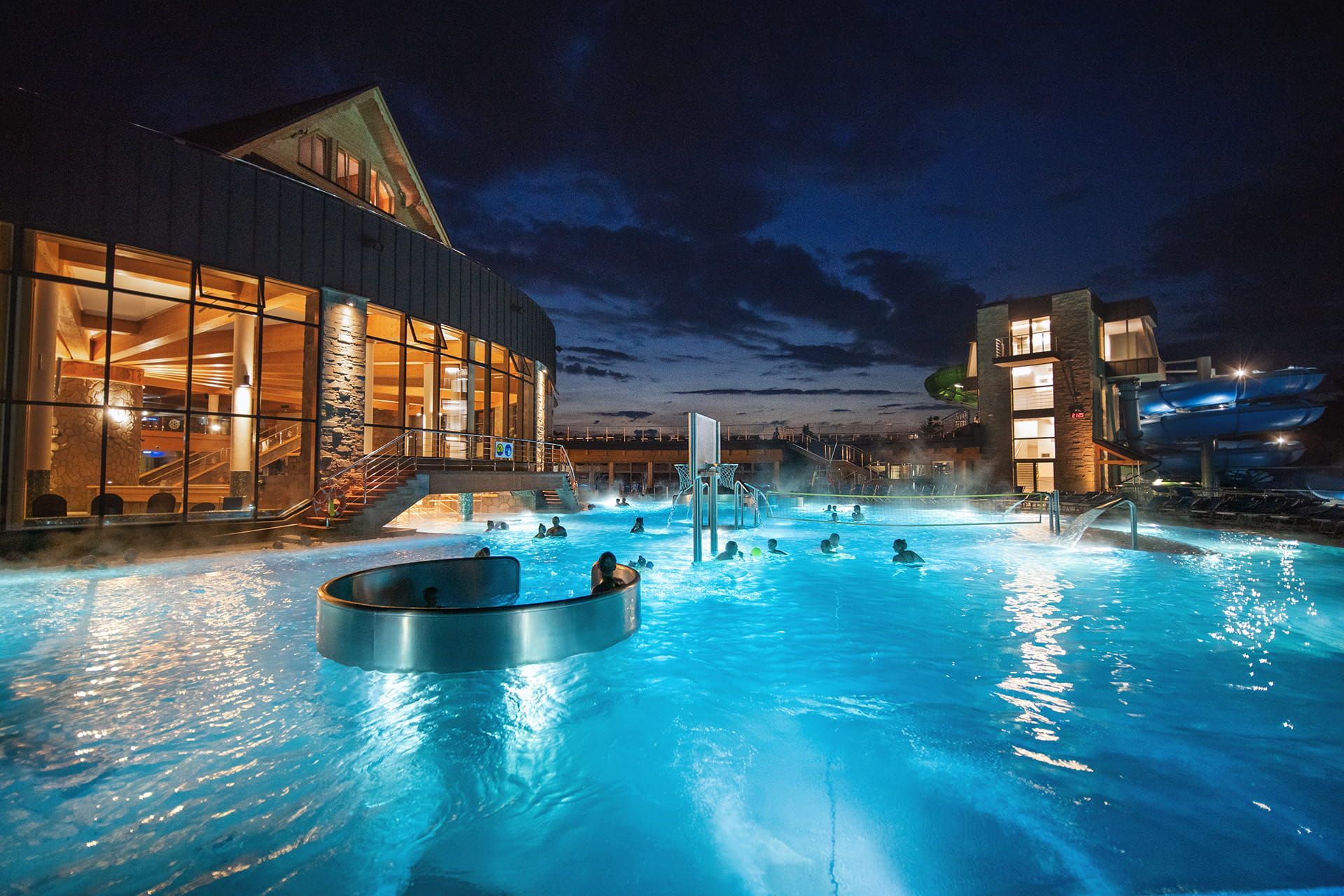 Guests in a steaming outdoor thermal pool at dusk beside the main building of Chochołowskie Termy
