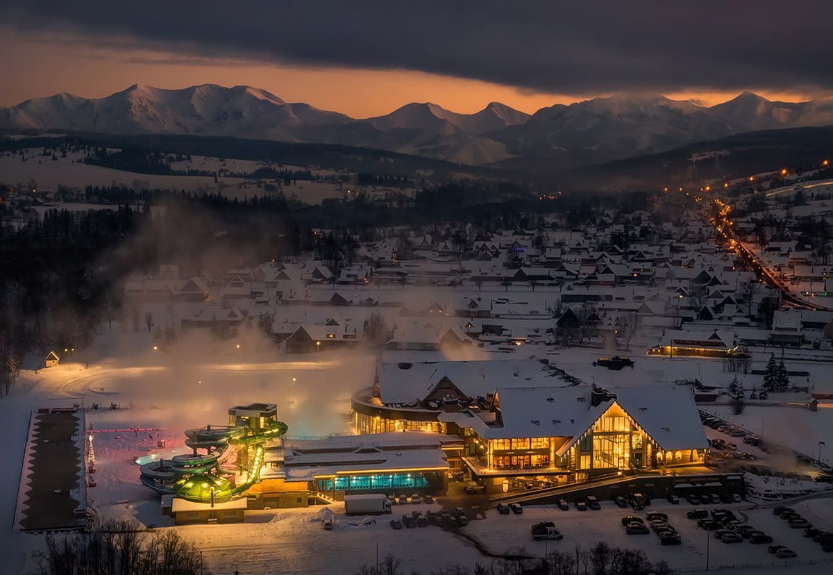 Aerial view of Chochołowskie Termy in winter, steam rising over the pools and the snowy Tatra Mountains at sunset
