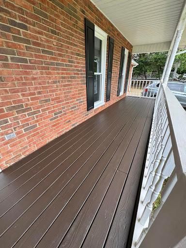 Covered porch with red brick wall, brown wood-look flooring, and white railing.