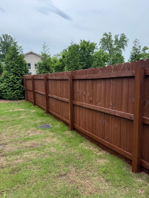 Brown wooden fence along a grassy yard with trees and a house in the background