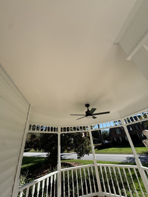 Covered porch with white railing, black ceiling fan, and view of green lawn and houses.