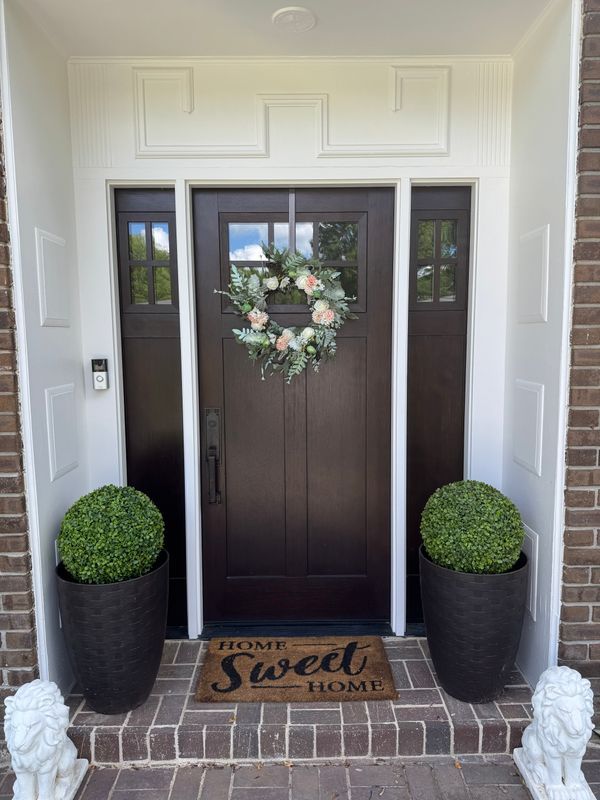 Front porch with dark brown door, wreath, black planters, and a welcome mat.
