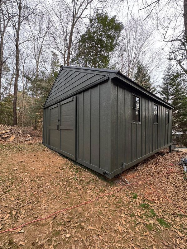 Dark gray metal shed with a slanted roof in a wooded yard in late fall.