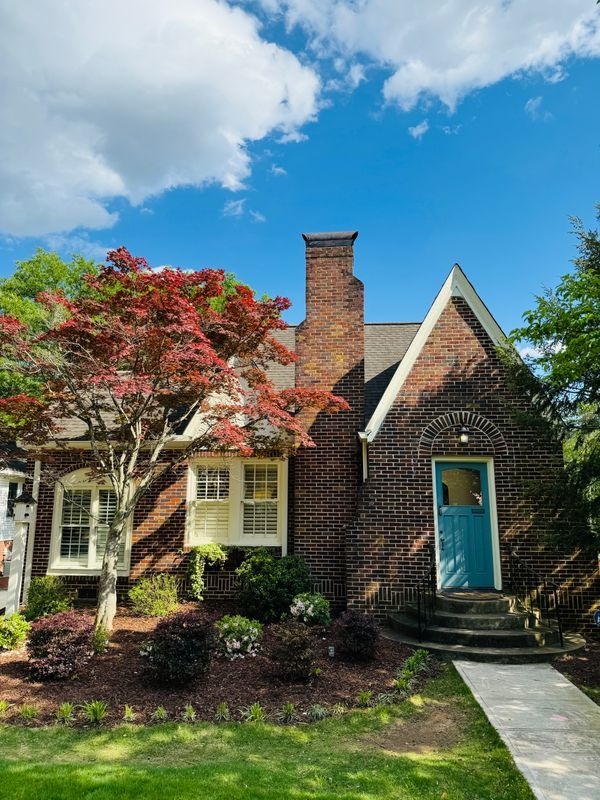 Brick cottage with blue door, white-trimmed windows, red-leaf tree, and walkway under a bright blue sky