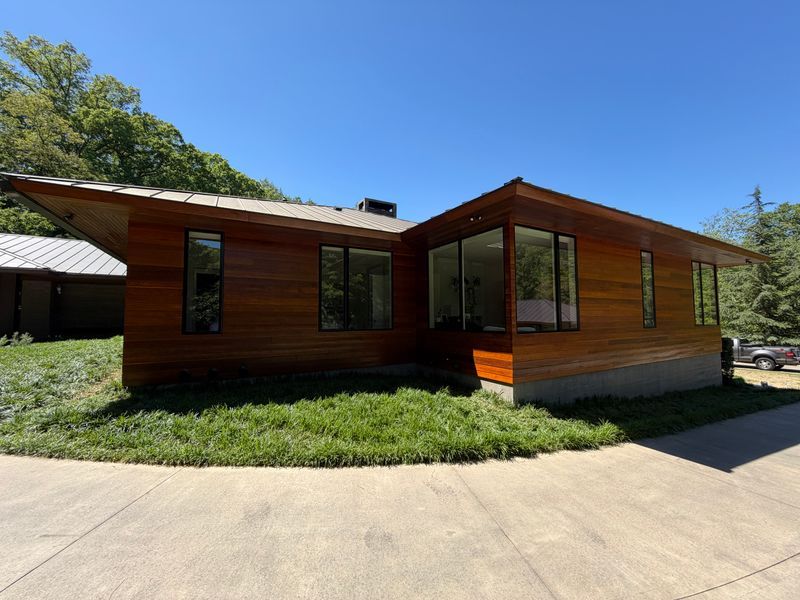 Modern single-story house with wood siding, large windows, and a curved concrete driveway in sunlight