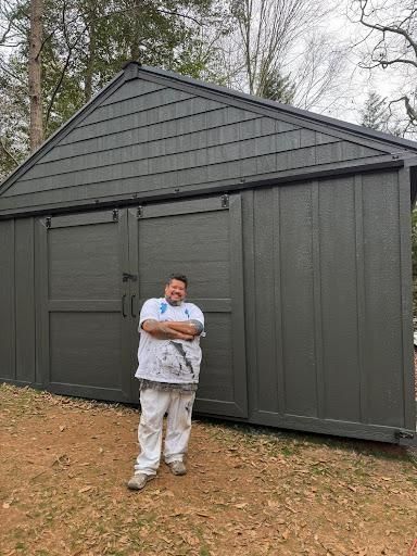 Person standing in front of a dark gray shed with arms crossed outdoors